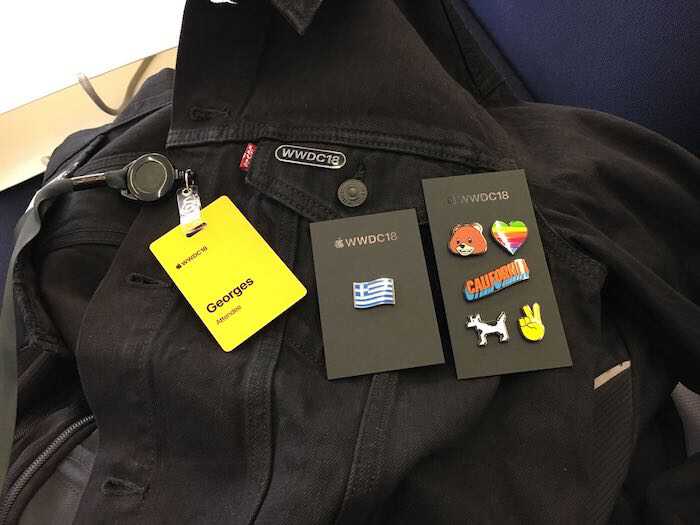 A photo of the WWDC18 black jacket with
									  layed out on top the yellow badge with
									  event's logo and the name George followed
									  by the word Attendee. There are two black
									  cards marked with AppleWWDC18 on top that
									  have colorful pins.  The first card has a
									  pin with the greek flag.  The second card
									  has six pins layed out in a rows of two
									  elements. The first row contains a bear
									  face and rainbow heart pin. The second row
									  contains the word California in a edged
									  design in orange and blue colors. The
									  third row contains the dogcow named Clarus
									  which is a bitmapped image designed by
									  Apple. The dogcow is black and white and
									  resembles a dog and a cow. The last pin is
									  a victory sign formed by the index and
									  middle fingers of a right hand.