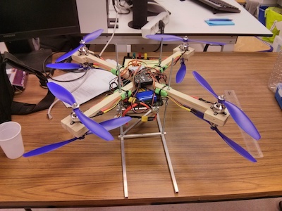 A photo of a handmade drone with a
										  wooden frame, with the controller and
										  cables exposed. The drone has a total
										  of eight fans/helices aranged in
										  inversed pairs. The drone is on a
										  wooden desktop.